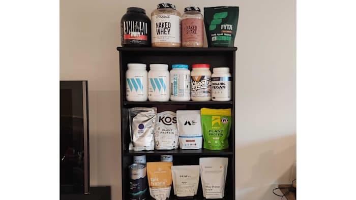 A four-shelf bookcase filled with various containers of protein powder.